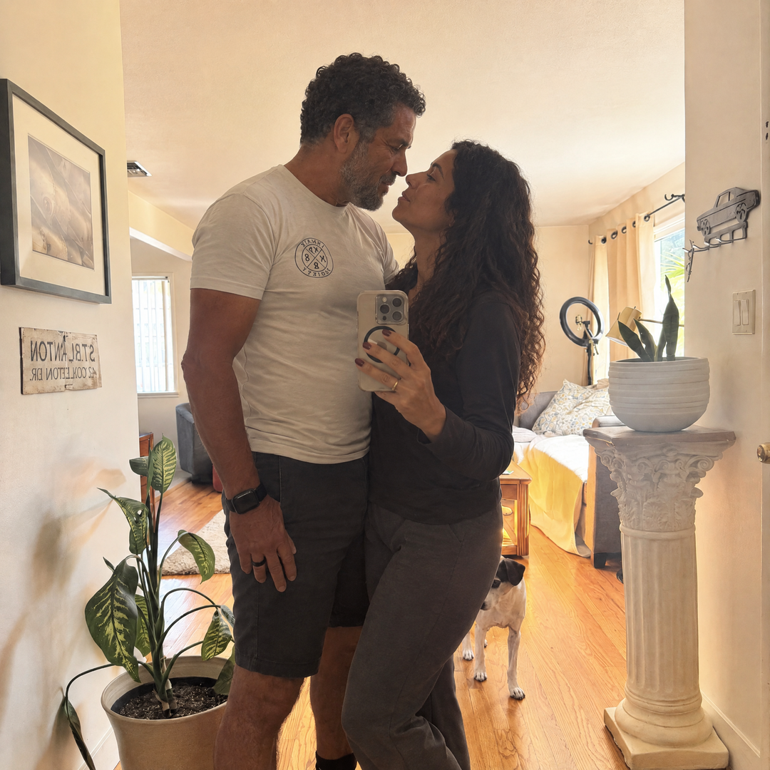 Casal tirando selfie no espelho com um cachorro ao fundo.
